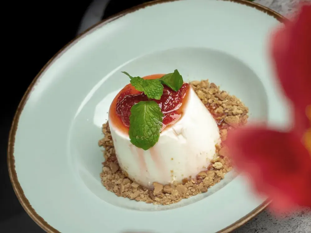 Panna cotta topped with strawberry compote and mint on a pale green plate, with a blurred red flower in the foreground.
