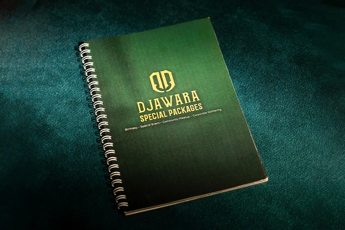 Spiral-bound Djawara special-packages menu booklet with gold lettering, photographed on deep green fabric.