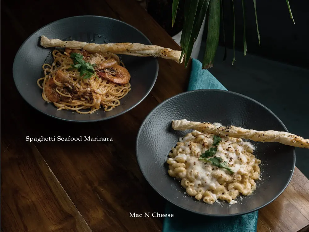 Overhead menu photo of two dark bowls labeled Spaghetti Seafood Marinara and Mac N Cheese on a wooden table.