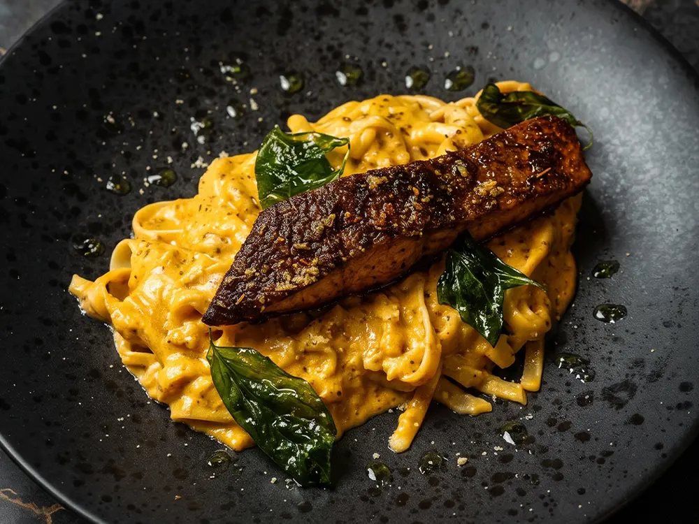Close-up of creamy fettuccine topped with a seared spiced fillet and crisp basil leaves on a textured black plate.