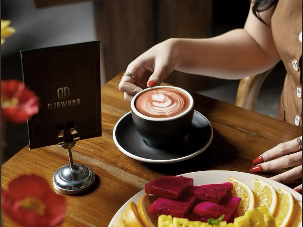 Café table scene with a hand reaching for a red velvet latte, a Djawara menu card, and a plate of dragon fruit and orange slices.