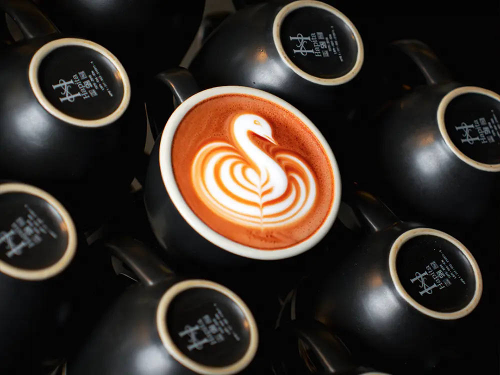 Overhead shot of matte black Hapita coffee cups surrounding one cup with detailed swan latte art.